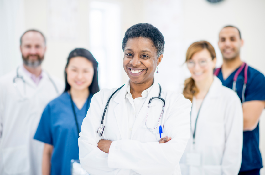 A diverse team of healthcare professionals, including a smiling doctor in a white coat and stethoscope, standing together in a clinical setting, conveying a sense of collaboration and care.